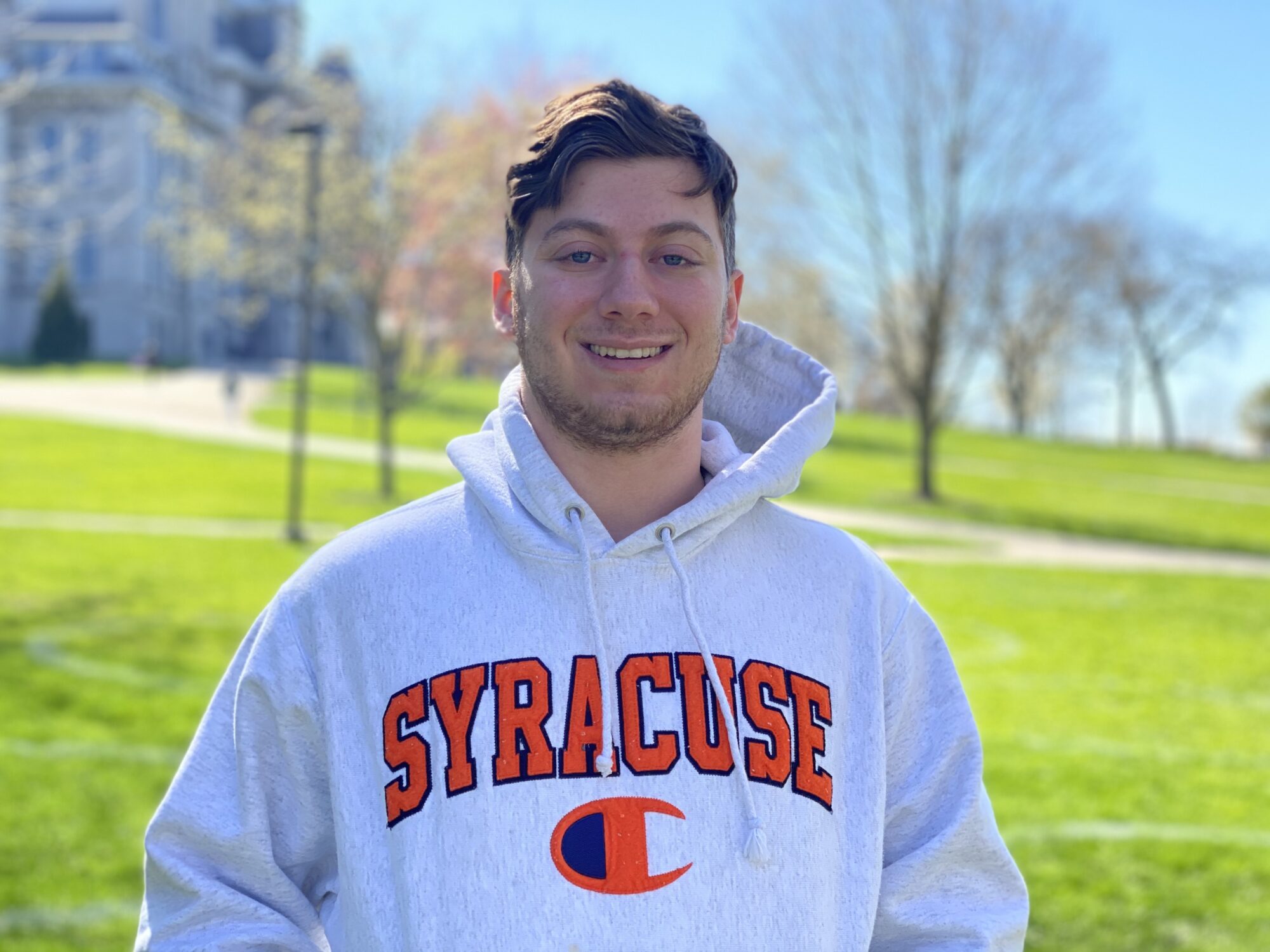 man wearing a Syracuse sweatshirt smiling in front of a grass field
