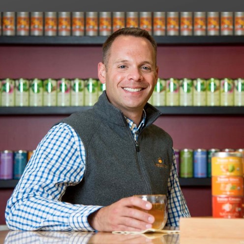 man holding a cup of tea in front of a wall of tea tins